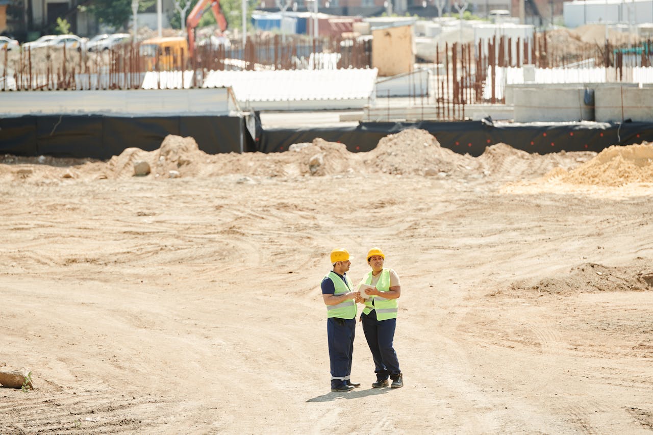services-03 Two workers discussing plans on a sandy construction site, wearing safety gear.